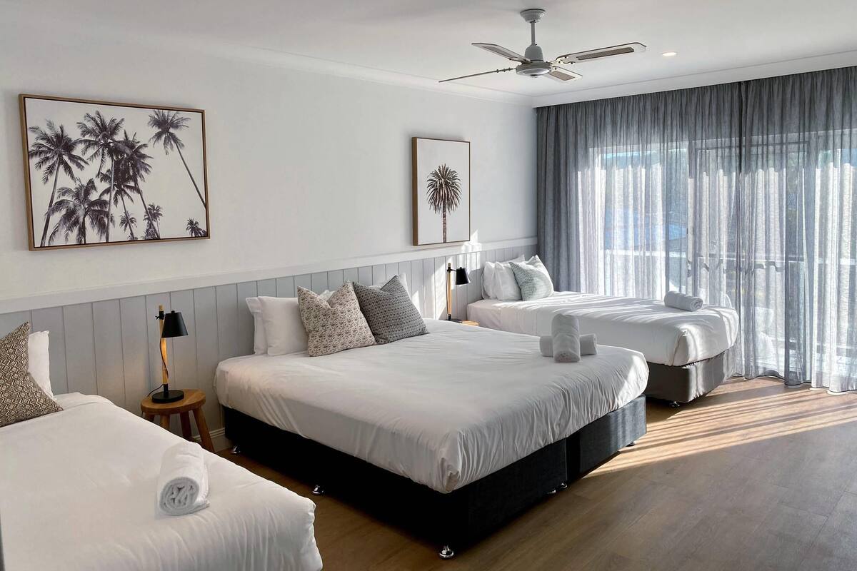 A hotel room with two beds, a ceiling fan, and large windows with sheer curtains.