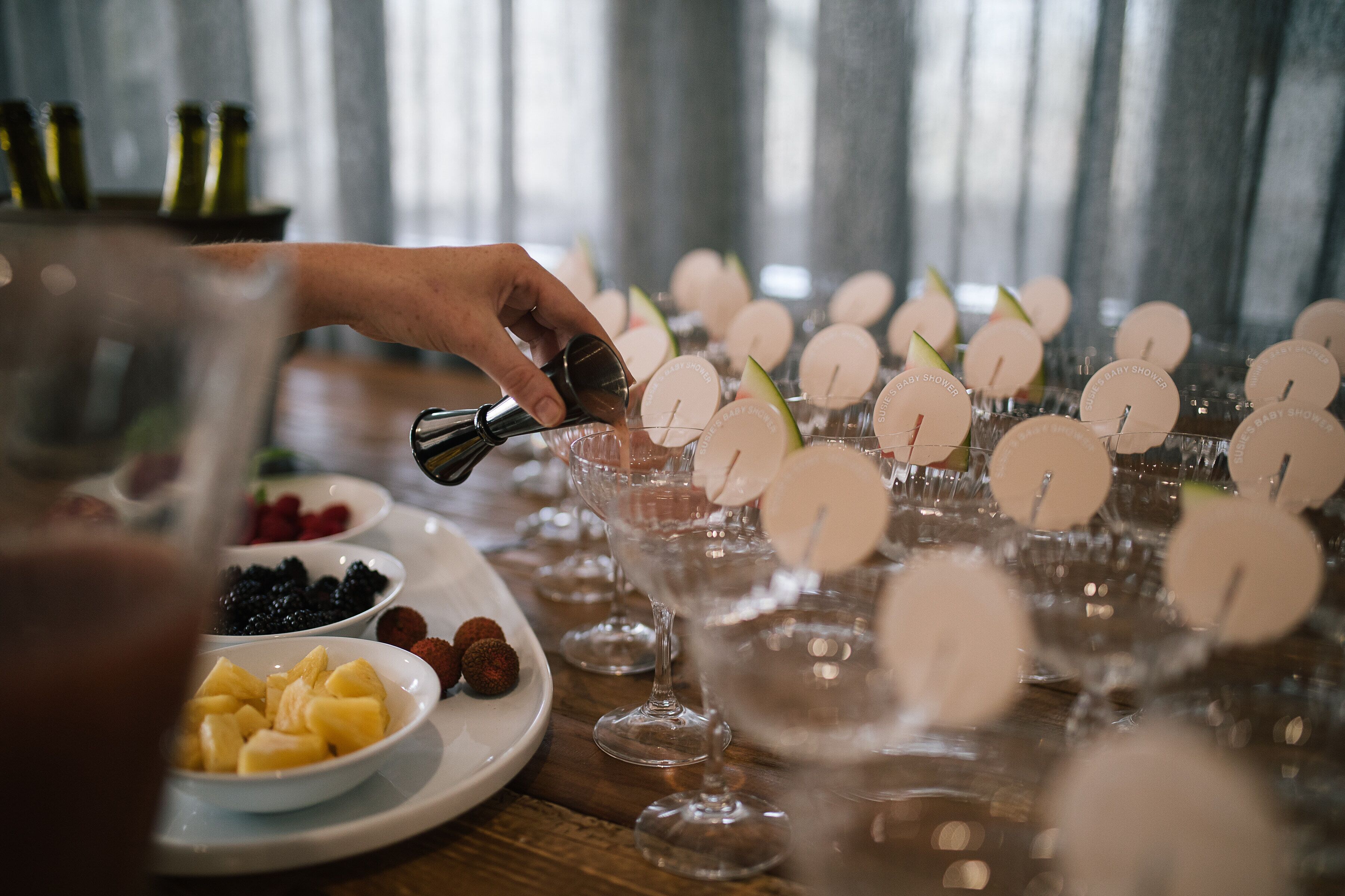 A table with multiple glasses, some filled with a clear liquid, and others with fruit slices. There are plates with various fruits and a bottle in the background.