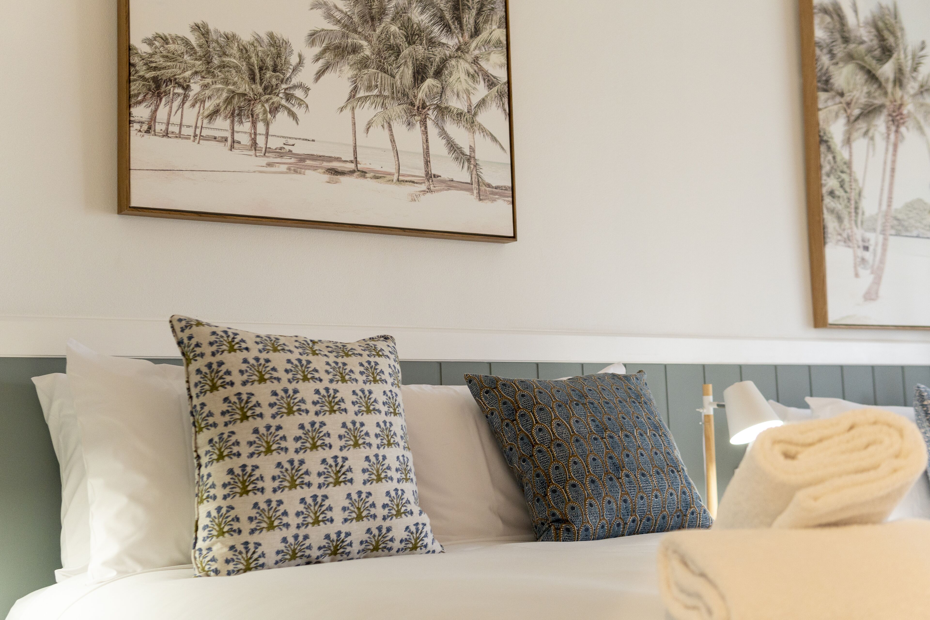 Una cama bien tendida con almohadones estampados, un cuadro con una playa en la pared y una toalla enrollada al lado.