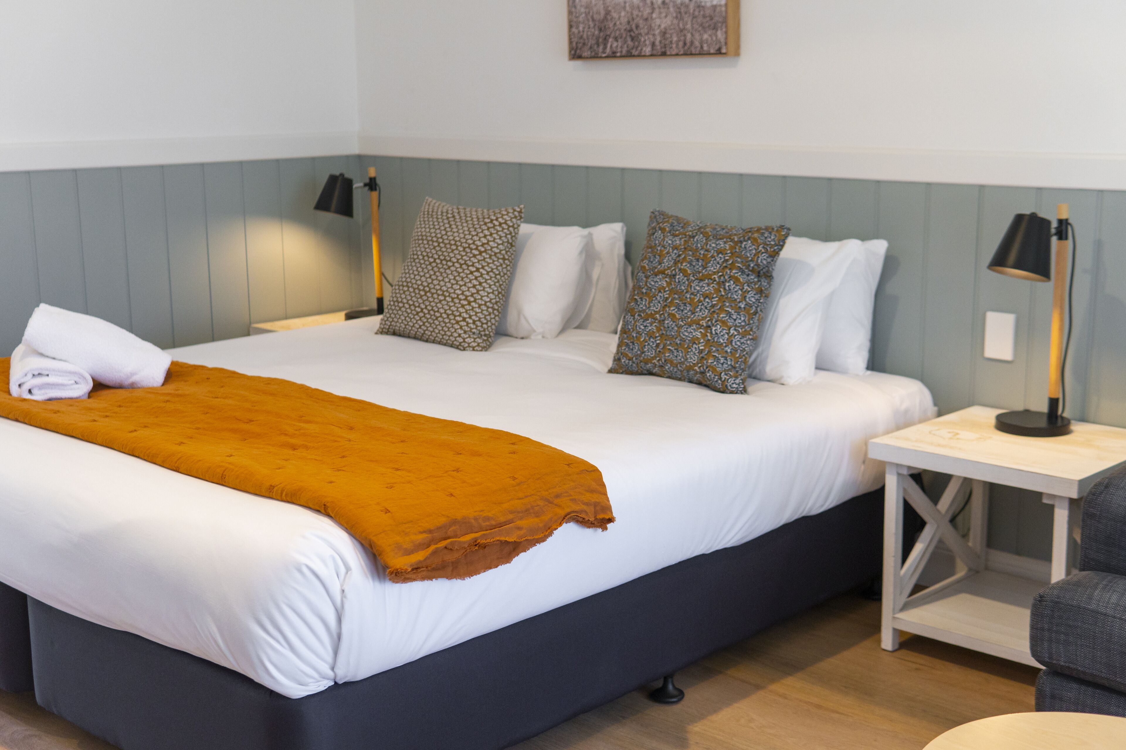A hotel room with a large bed, a nightstand, a chair, and a wall-mounted lamp.