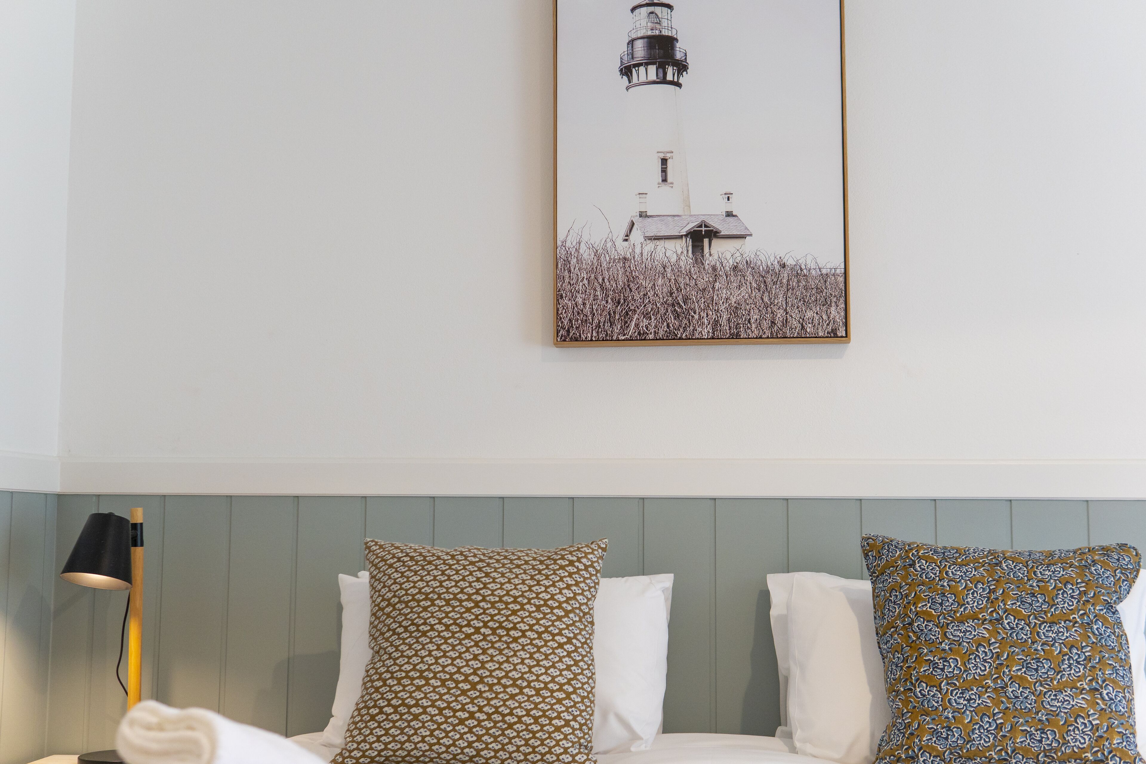 Une chambre d’hôtel avec un lit, une table de chevet, une lampe et un tableau représentant un phare.