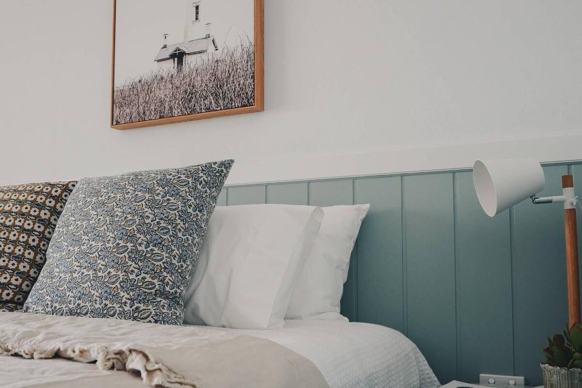 A bedroom with a bed, patterned pillows, a white pillow, a lamp, a magazine, and a framed picture on the wall.