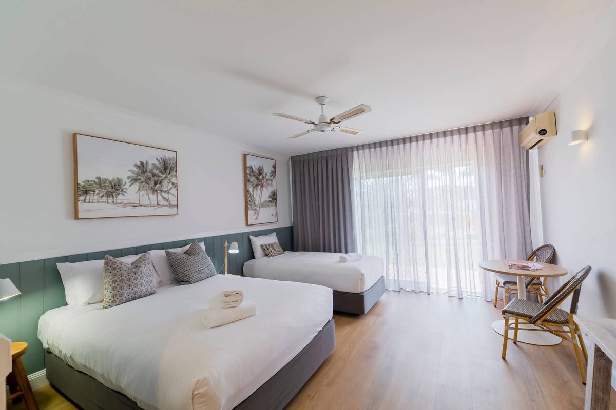 A hotel room with two beds, a ceiling fan, a table with chairs, and a window with curtains.