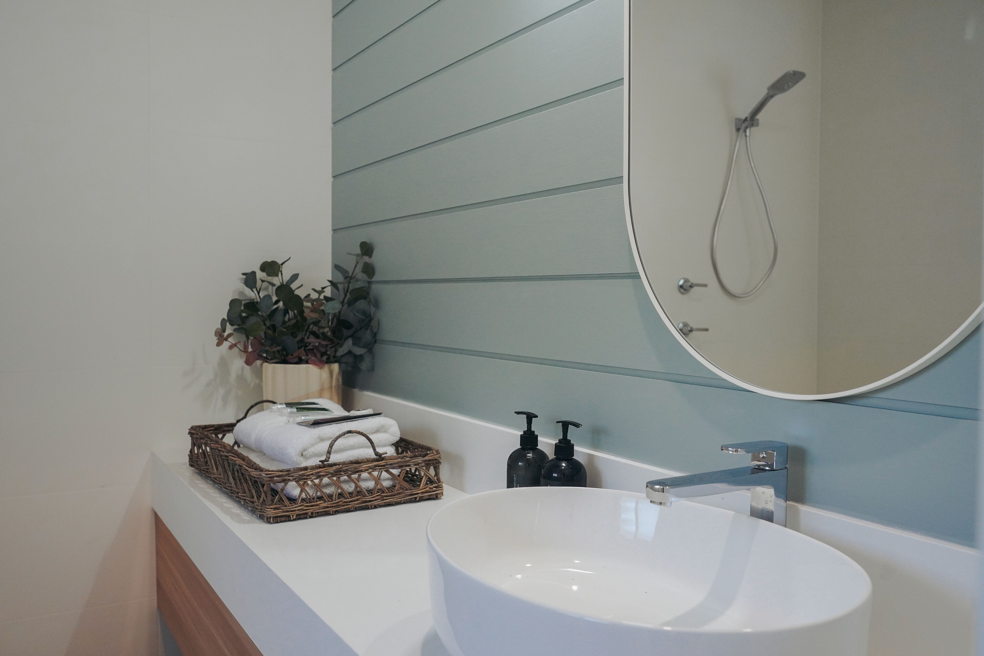 A bathroom with a white sink, a mirror, a shower, and a wicker basket with towels.