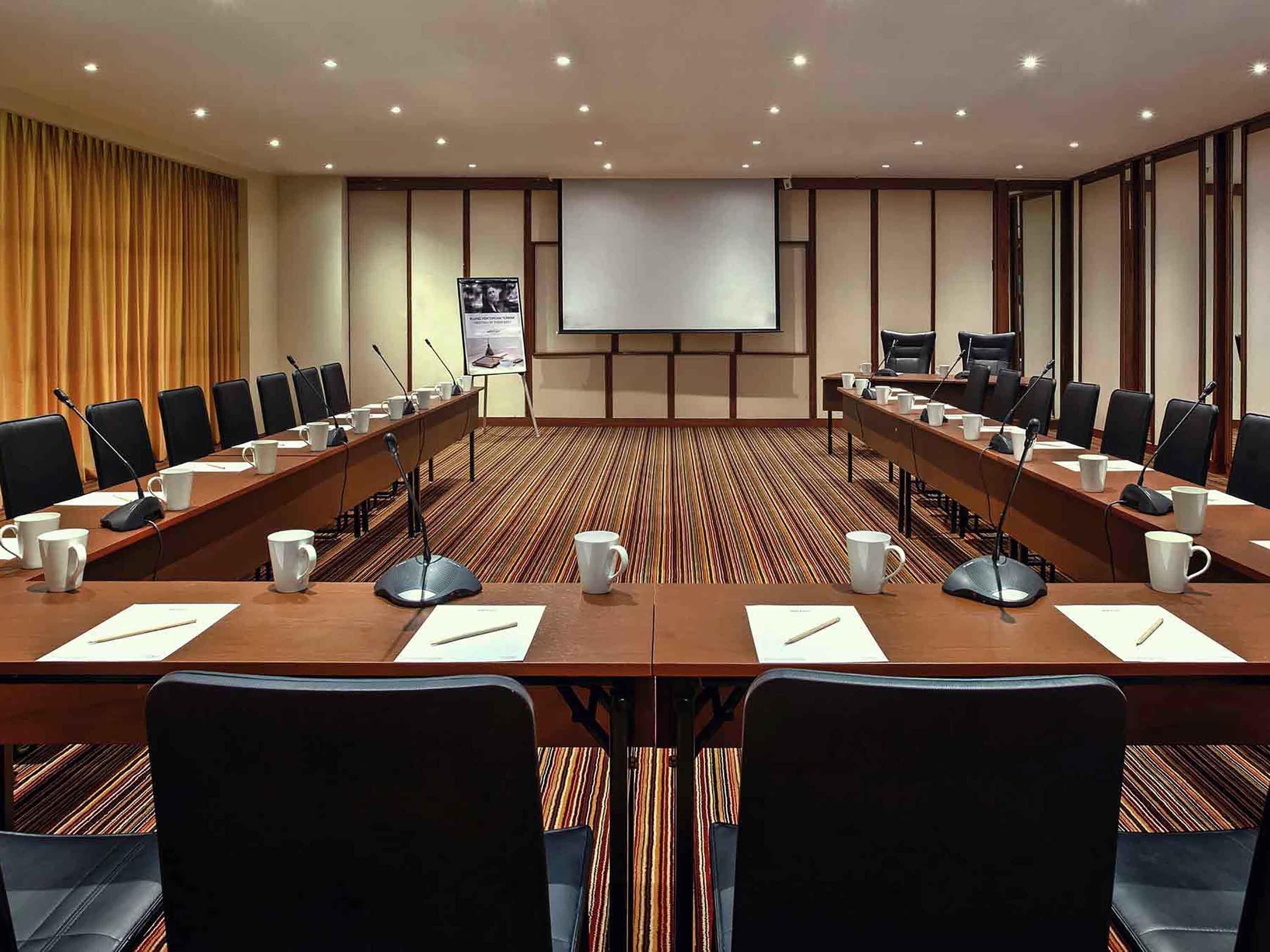 Una sala de conferencias con una larga mesa, varias sillas, micrófonos y una pantalla de proyector.
