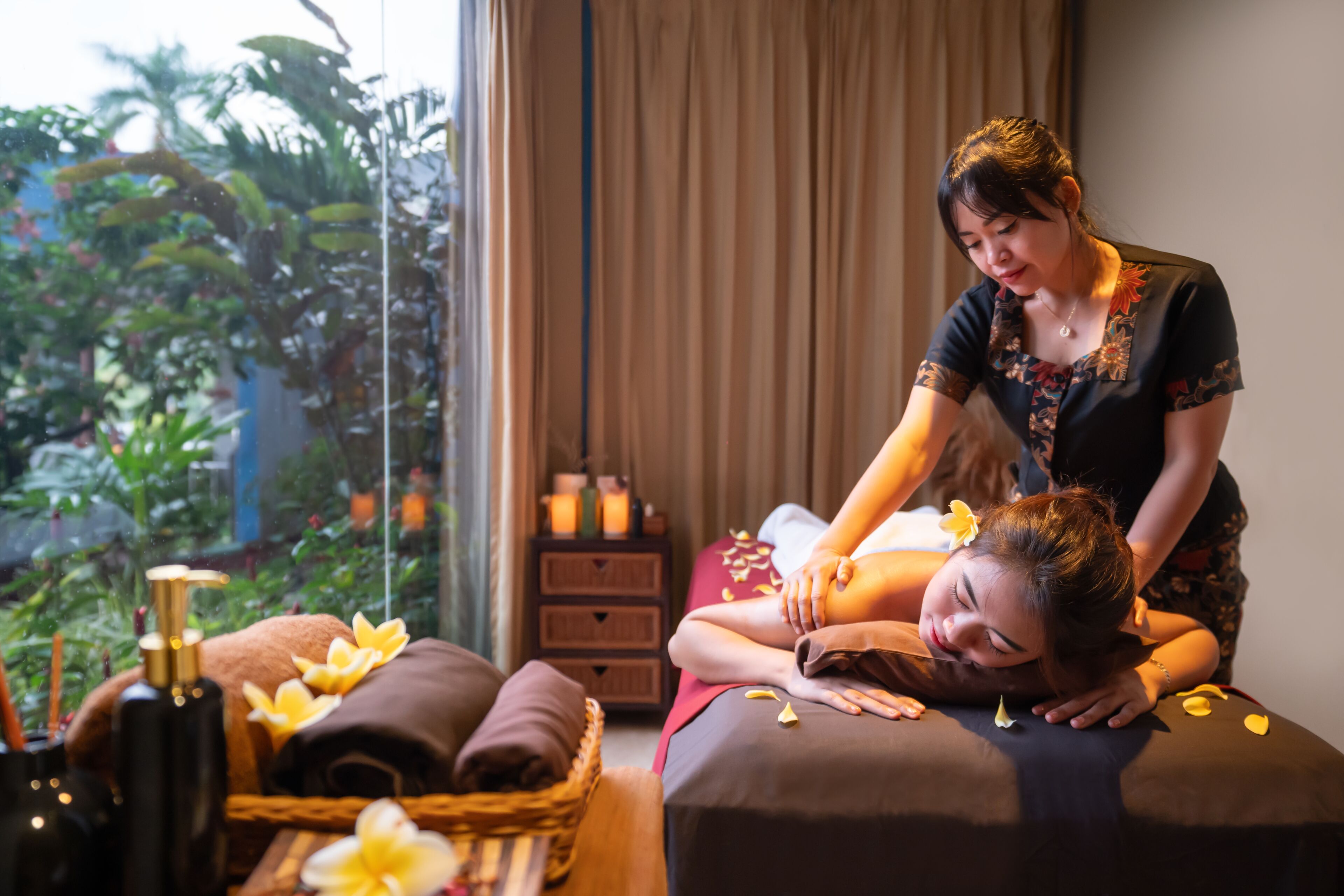 A massage therapy session in a room with a view of greenery outside, a massage table, and a wooden side table with towels and a bottle.