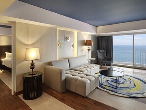 Une chambre d’hôtel moderne avec un canapé, une table basse ronde, un fauteuil, une lampe et une vue sur la mer.