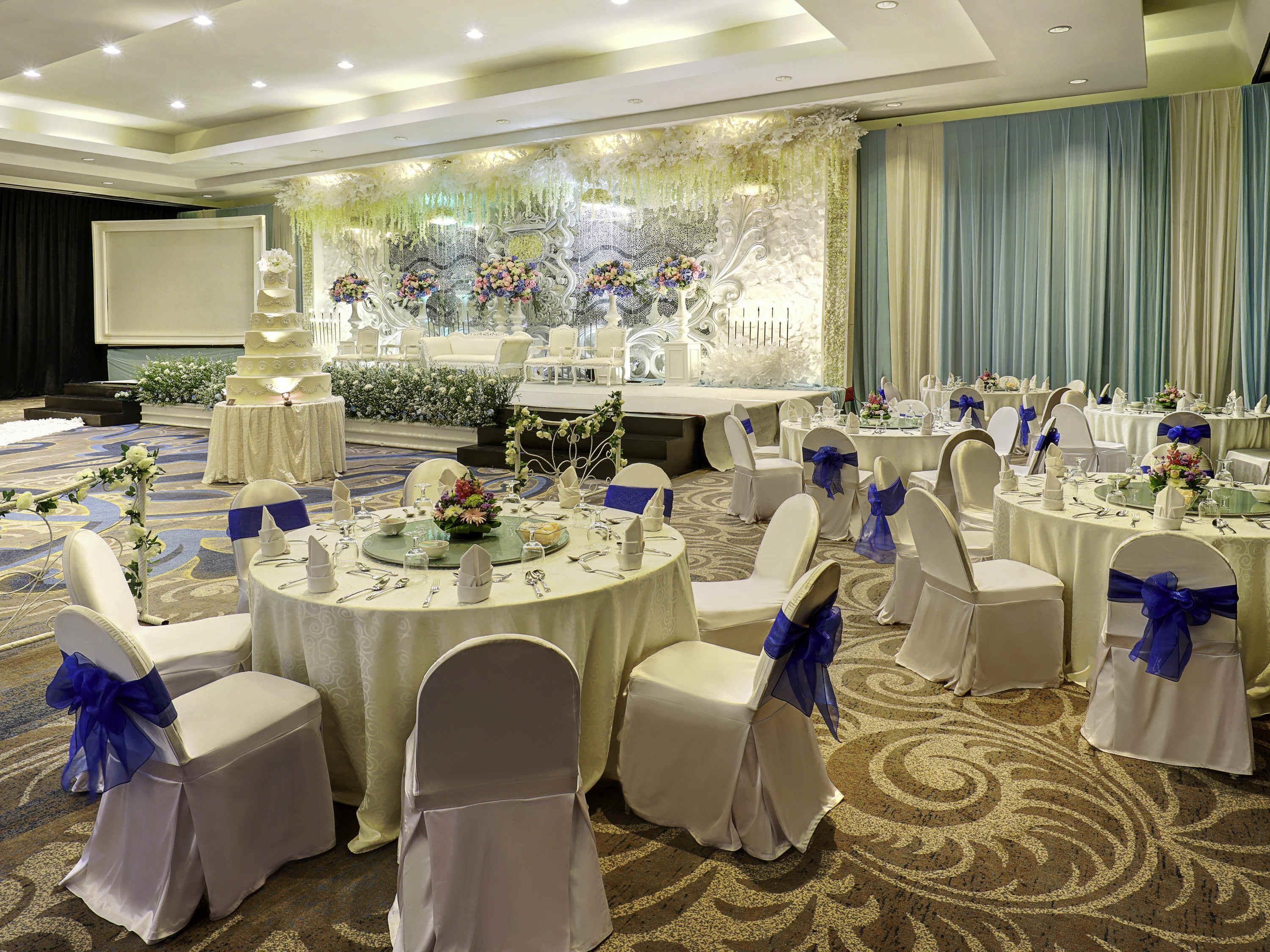 A banquet hall with round tables, white chairs with blue sashes, and floral centerpieces.