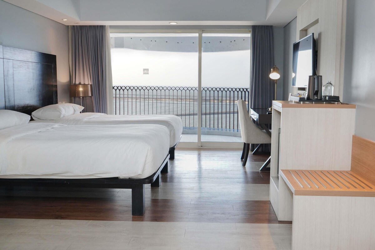 A hotel room with a large bed, a desk, a chair, a TV, and a balcony with a view of the sea.