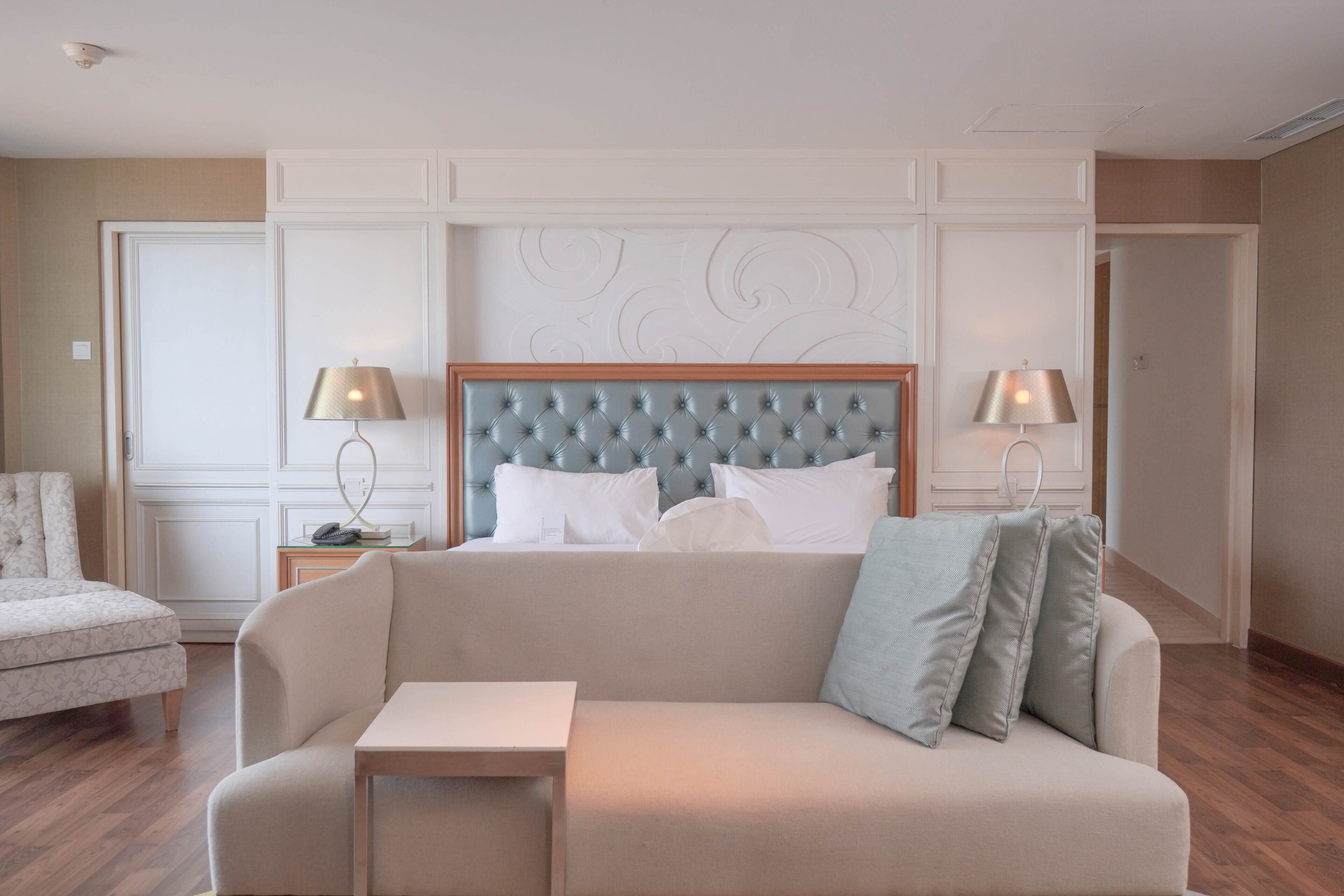 Habitación de hotel con cabecera capitonada, un sofá, una mesita y dos lámparas de noche.