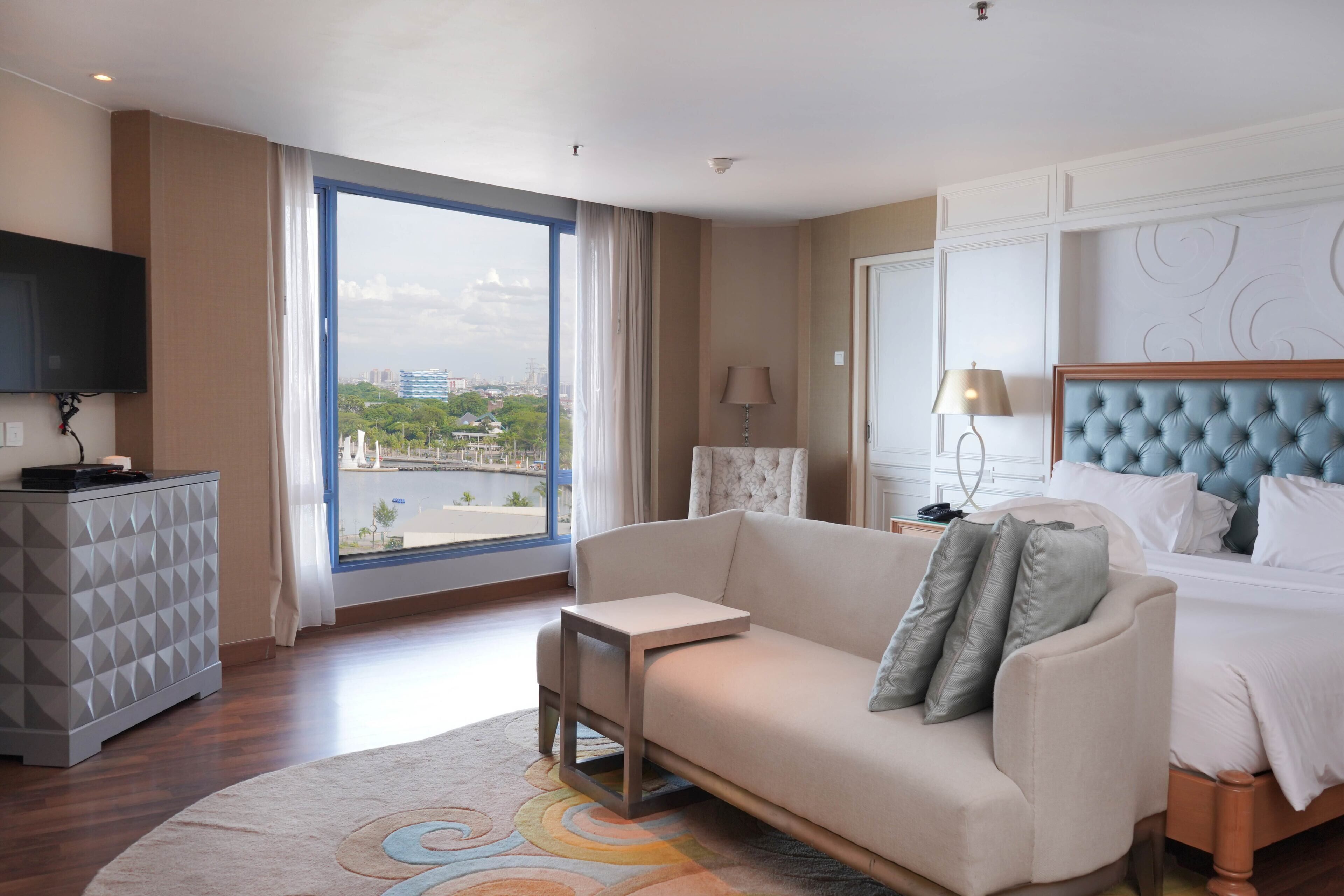 A hotel room with a large window, a bed with a blue headboard, a sofa, a small table, and a TV on a cabinet.
