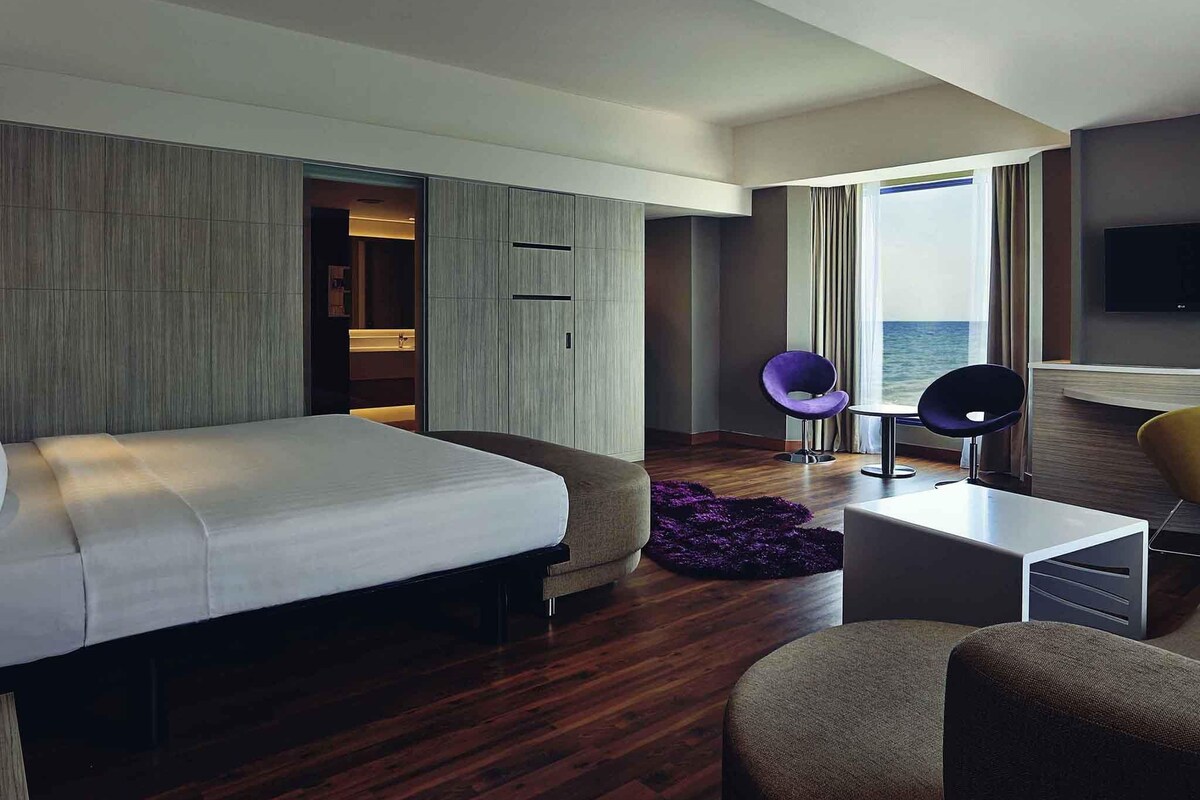 A modern hotel room with a large bed, a small table, two chairs, a sofa, and a view of the sea.