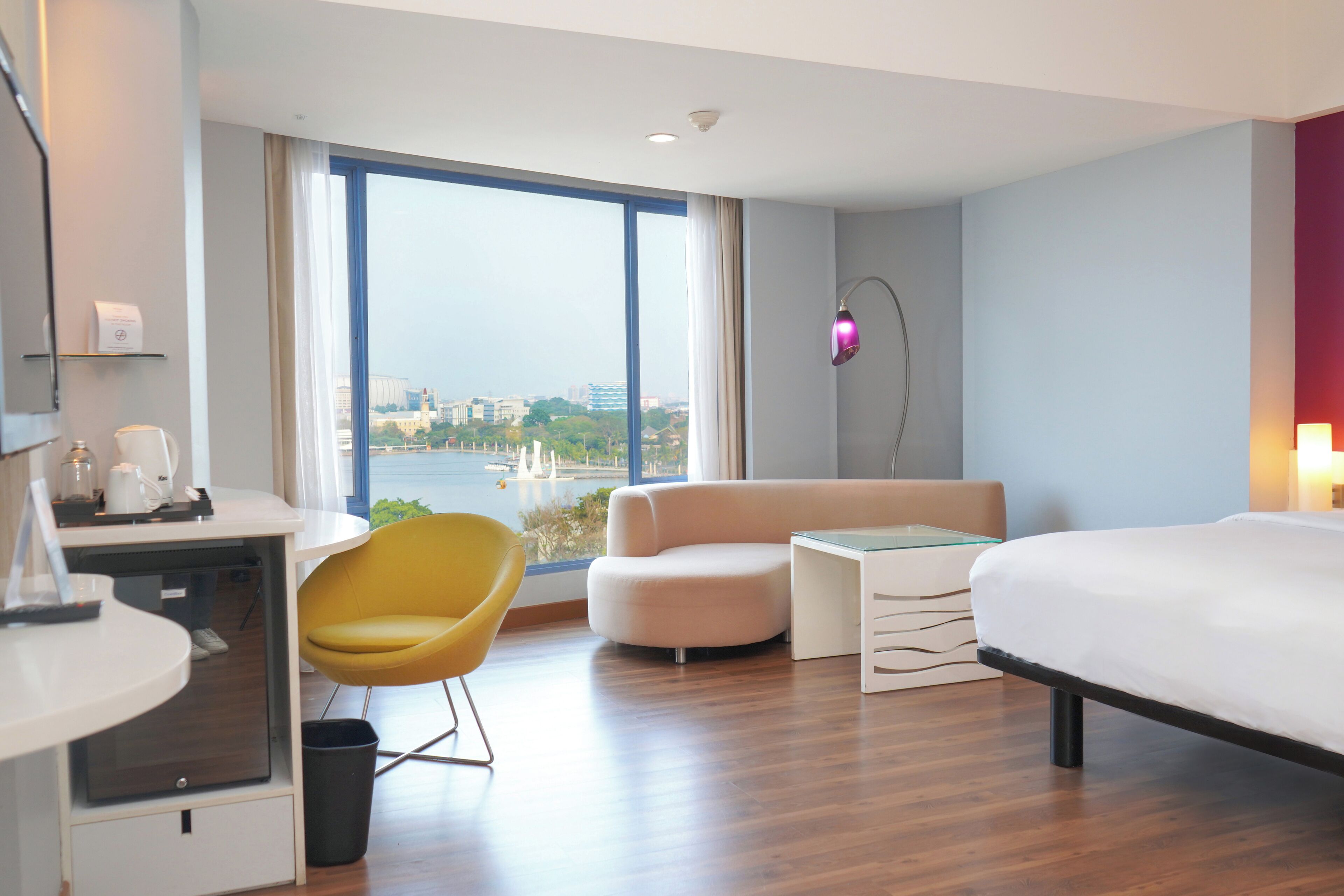 A modern hotel room with a large window, a bed, a desk, a chair, and a small table.