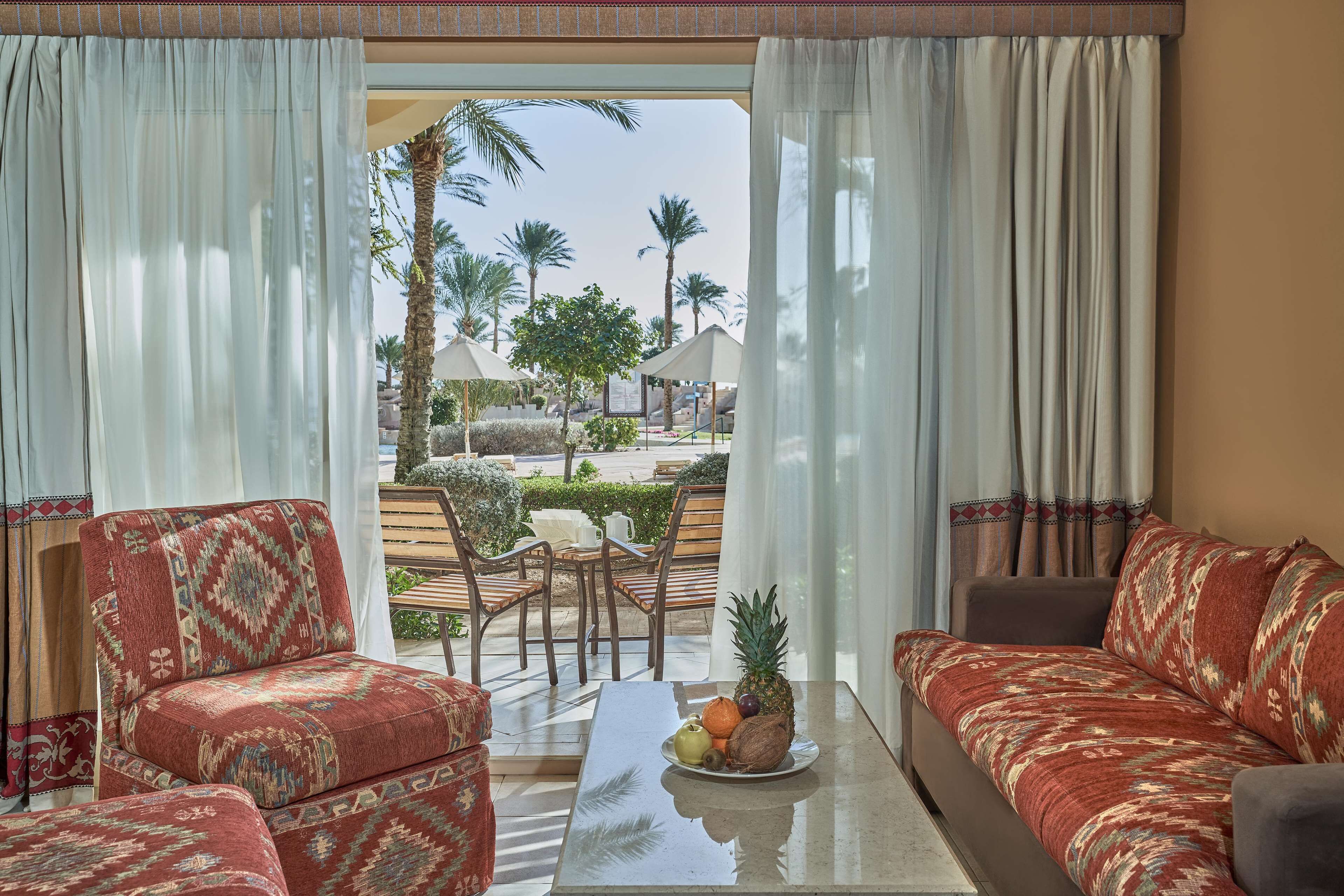 A room with a sofa and armchairs, a table with fruit, and a view of palm trees and a pool.