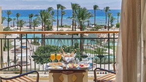 Ein Balkon mit Frühstückstisch, mit Blick auf einen Pool und Strand, vorbei an Palmen und Bergen in der Ferne.