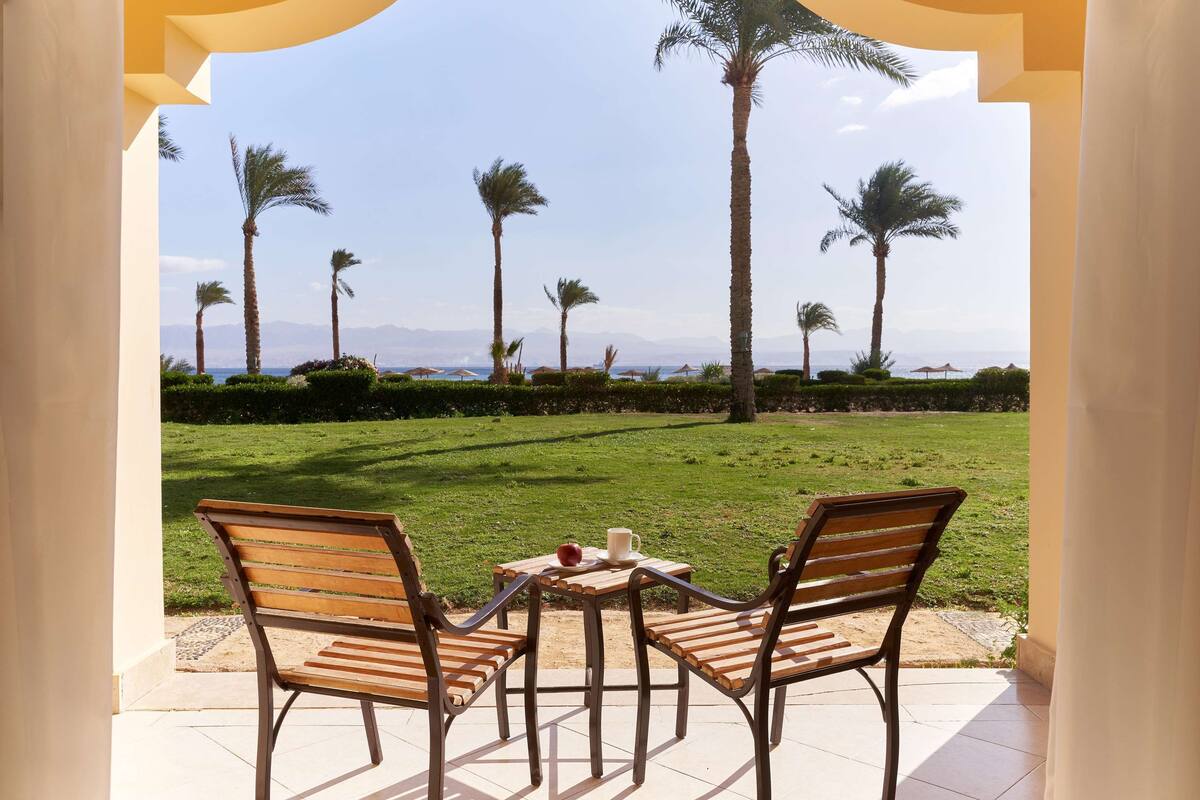 Een terras met twee houten stoelen en een kleine tafel, met uitzicht op een gazon met palmbomen en een verre bergweergave.