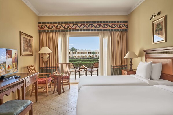 Une chambre d’hôtel avec un grand lit, un bureau avec un ordinateur, une chaise, une petite table et une vue sur un immeuble et la verdure environnante.