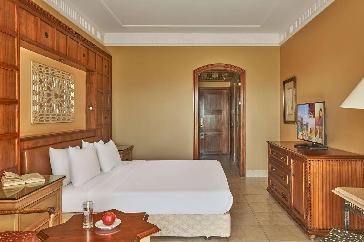 Een hotelkamer met een groot bed, een houten hoofdbord, een bureau met een lamp, een televisie en een decoratief wandobject.