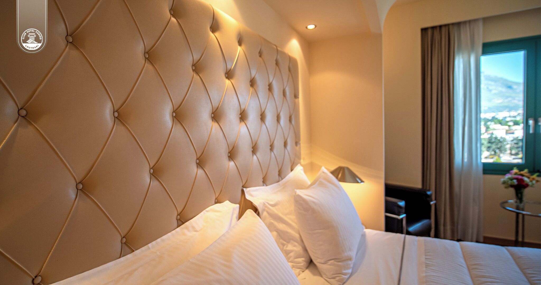 Une chambre d’hôtel avec une tête de lit capitonnée, du linge de lit blanc, une petite table avec une lampe et une vue sur l’extérieur par une fenêtre.
