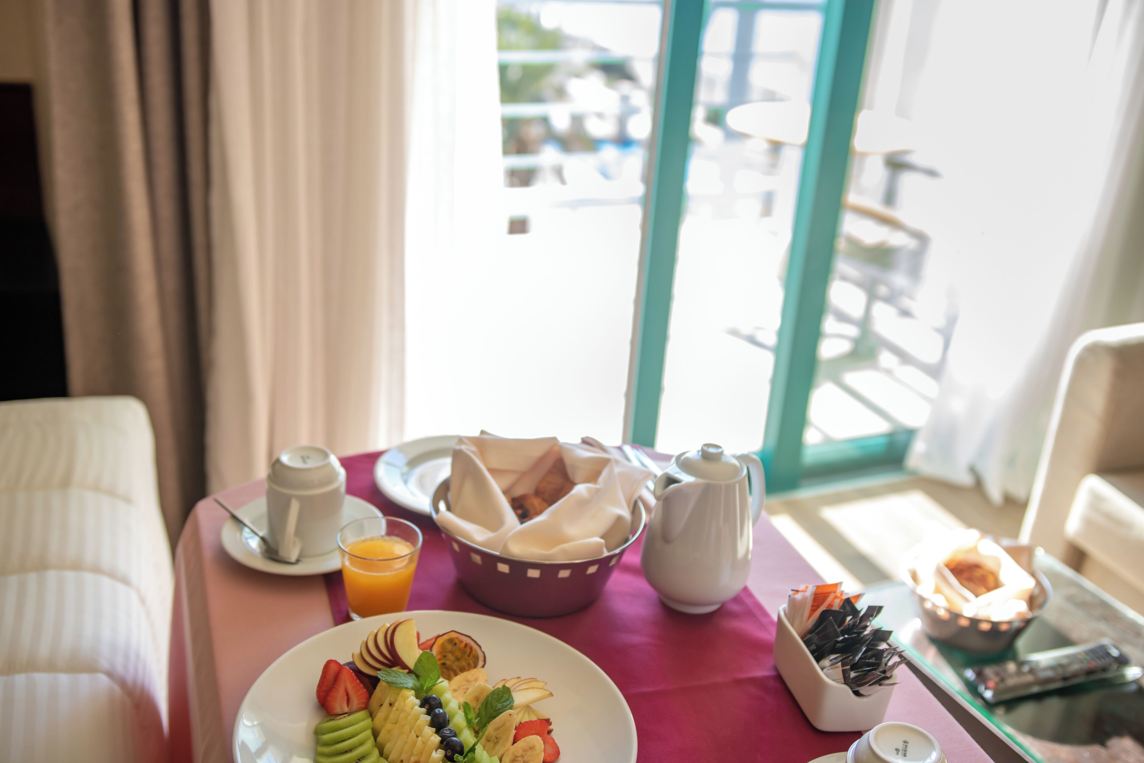 Una mesa de desayuno con una bandeja de frutas, un vaso de jugo de naranja, un calentador de agua, un cuenco de servilletas y un control remoto sobre una mesa de vidrio.