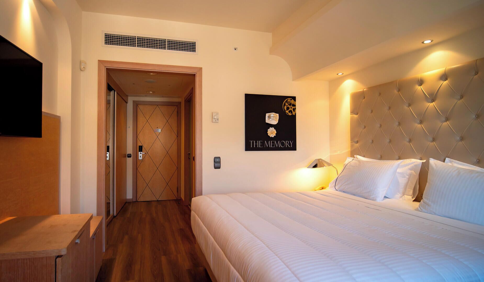 A hotel room with a bed, a bedside table, a wall-mounted TV, a framed picture, and a door with a sign.