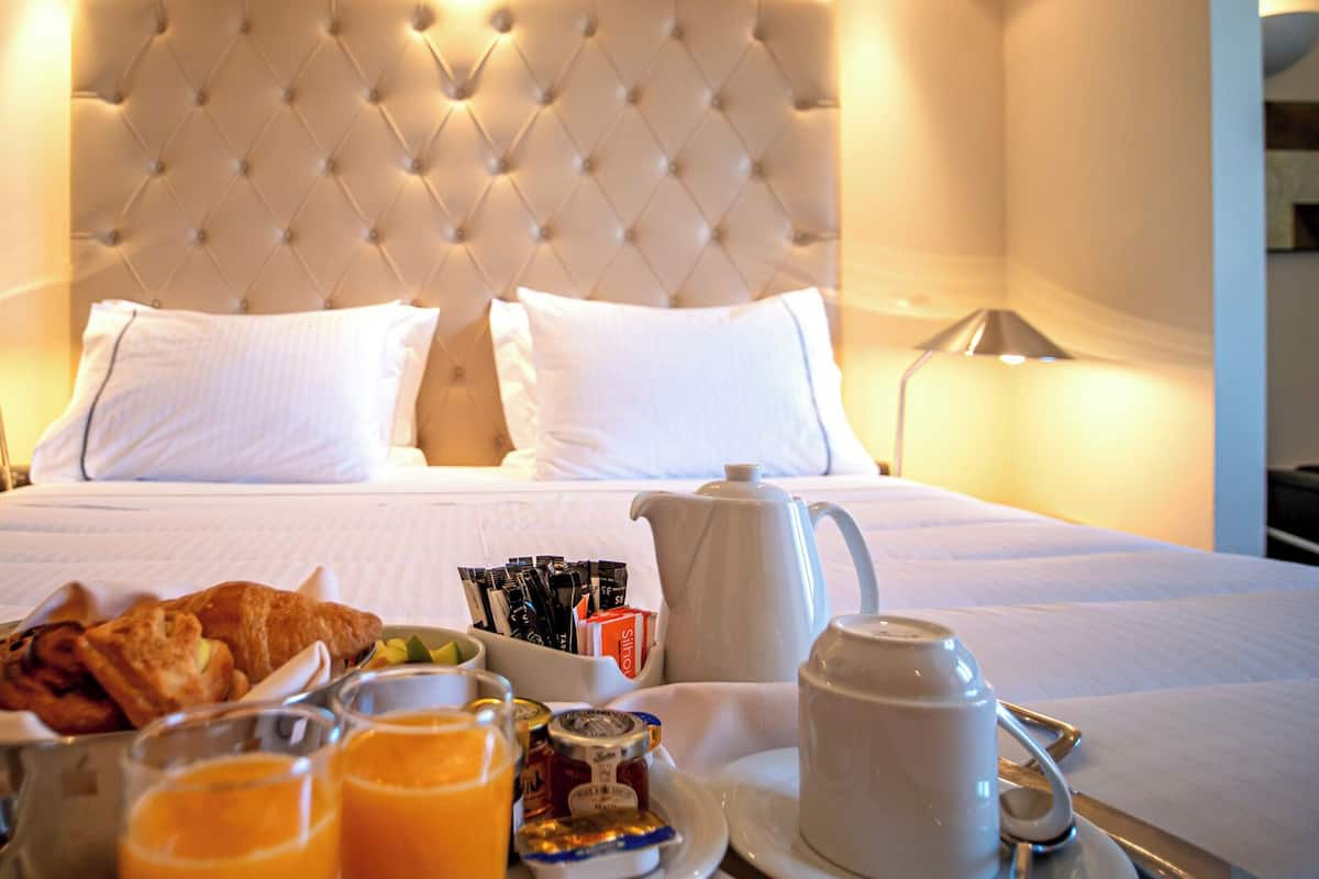 Ein Hotelzimmer mit einem großen Bett, weißen Leinen, einem gepolsterten Kopfteil, Nachttischlampen und einem Frühstückswagen mit Croissants, Saft und Beilagen.