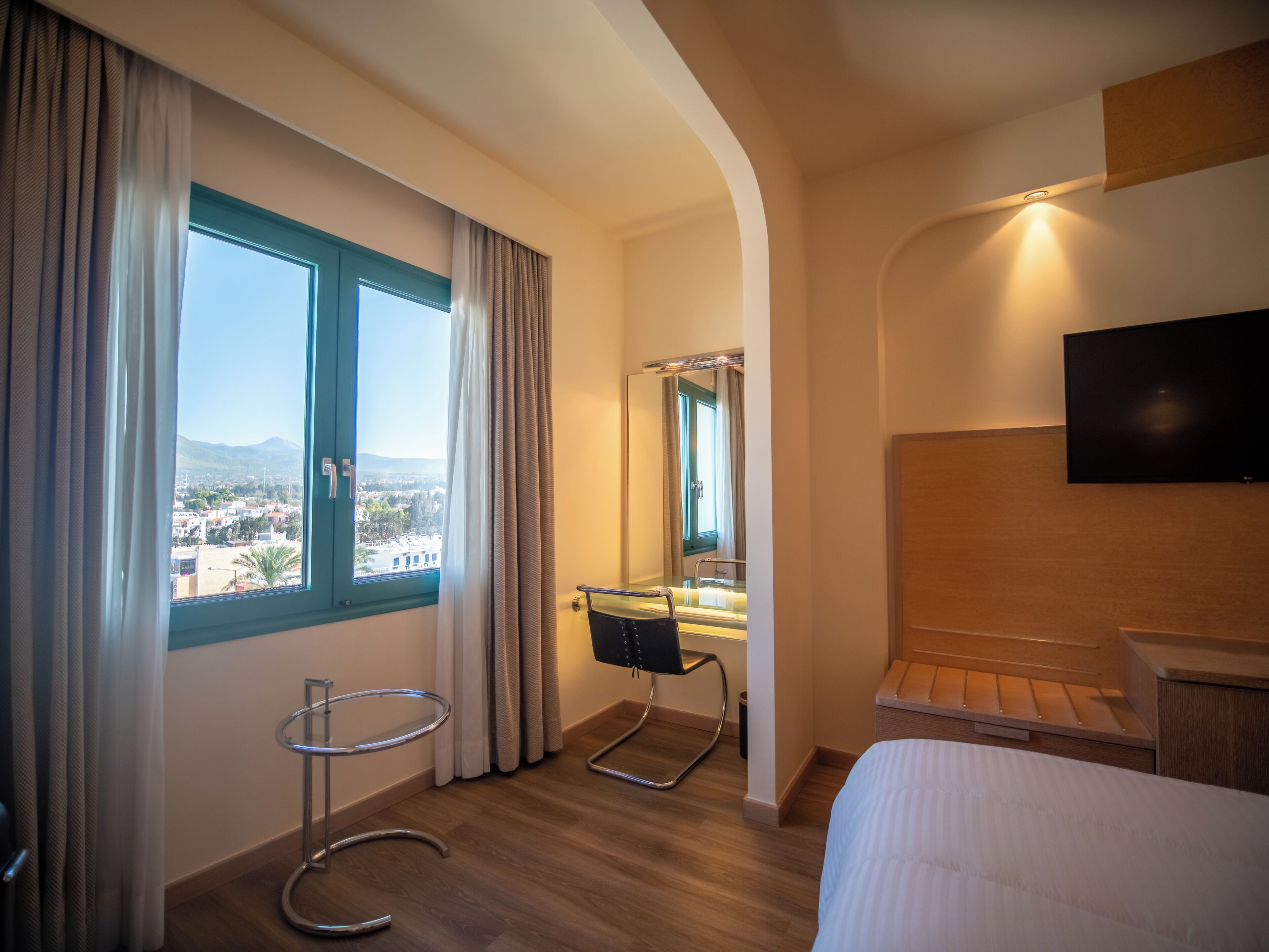 A hotel room with a bed, a TV, a mirror, a chair, and a window with curtains.