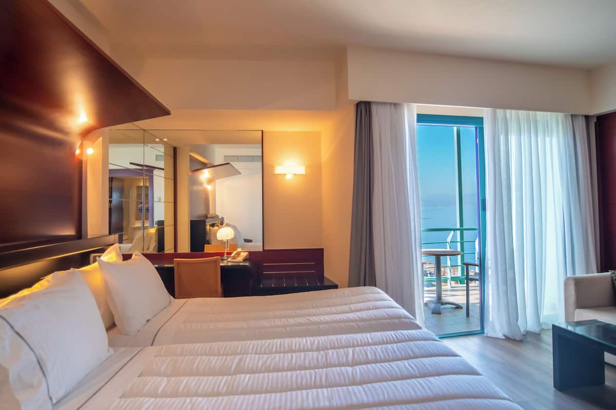 Ein Hotelzimmer mit einem großen Bett, einem Schreibtisch mit Lampe, einem Sessel, einem kleinen Tisch und einem Balkon mit Blick.