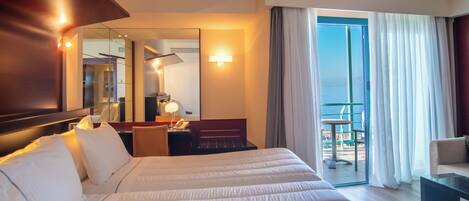 A hotel room with a large bed, a desk with a lamp, a chair, a small table, and a balcony with a view.
