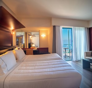 Quarto de hotel com uma cama grande, uma escrivaninha com luminária, uma cadeira, uma mesa pequena e uma varanda com vista.