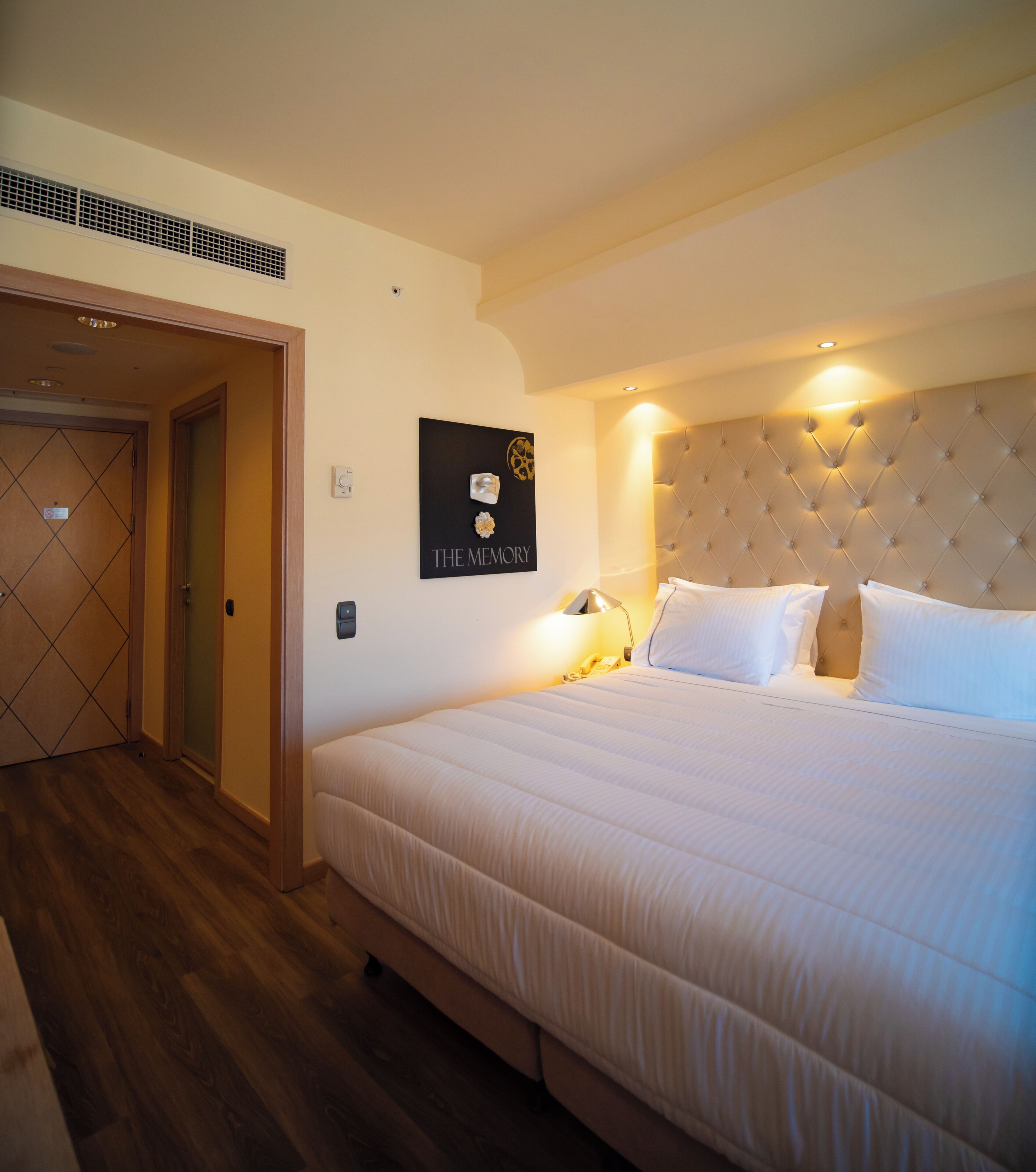 Habitación de hotel con una cama grande, un cabecero con diseño acolchado, una obra de arte en la pared titulada 'THE MEMORY' y una puerta con un patrón geométrico.