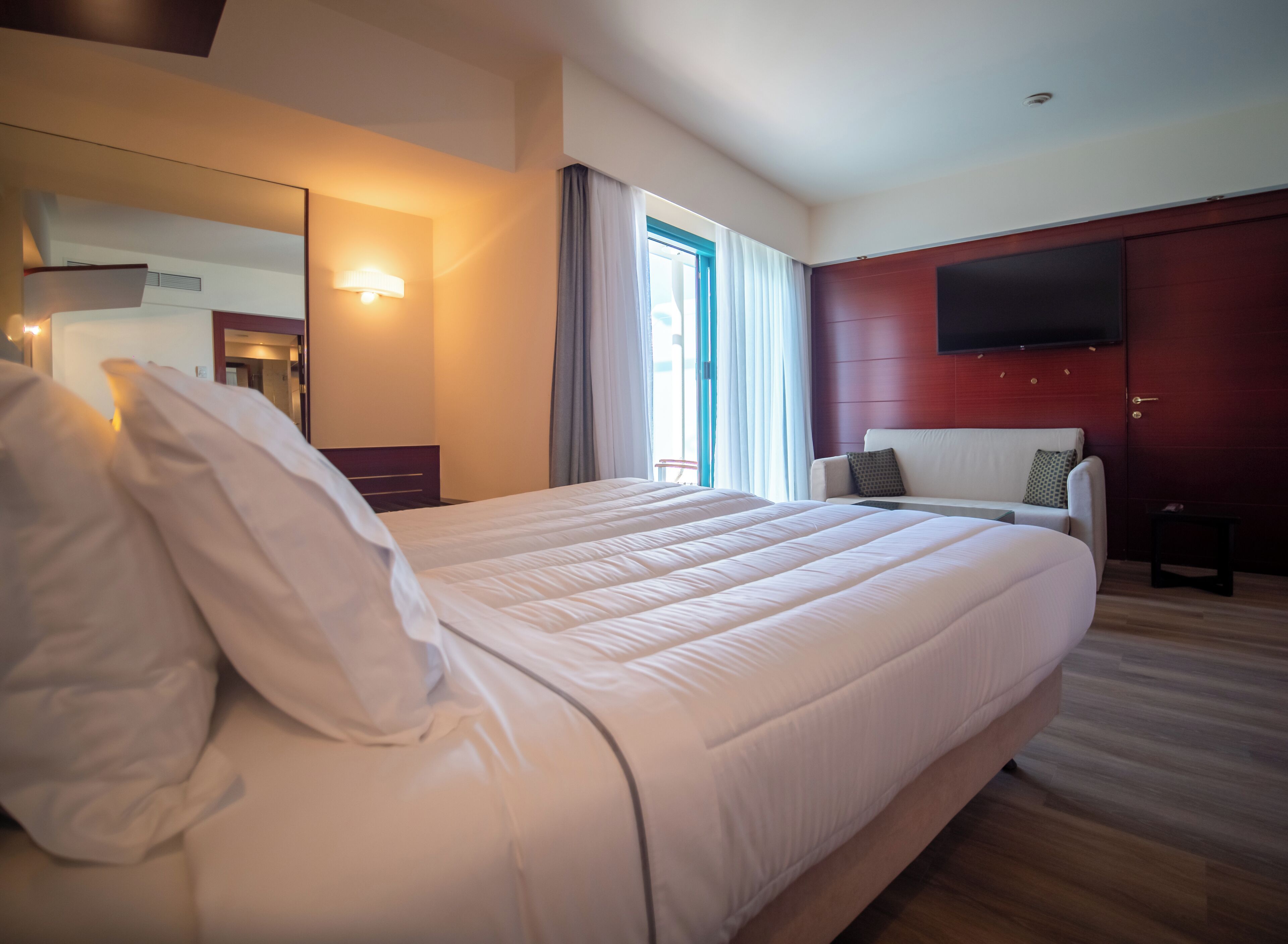 Quarto de hotel com uma cama grande, sofá, televisão na parede e janela com cortinas.