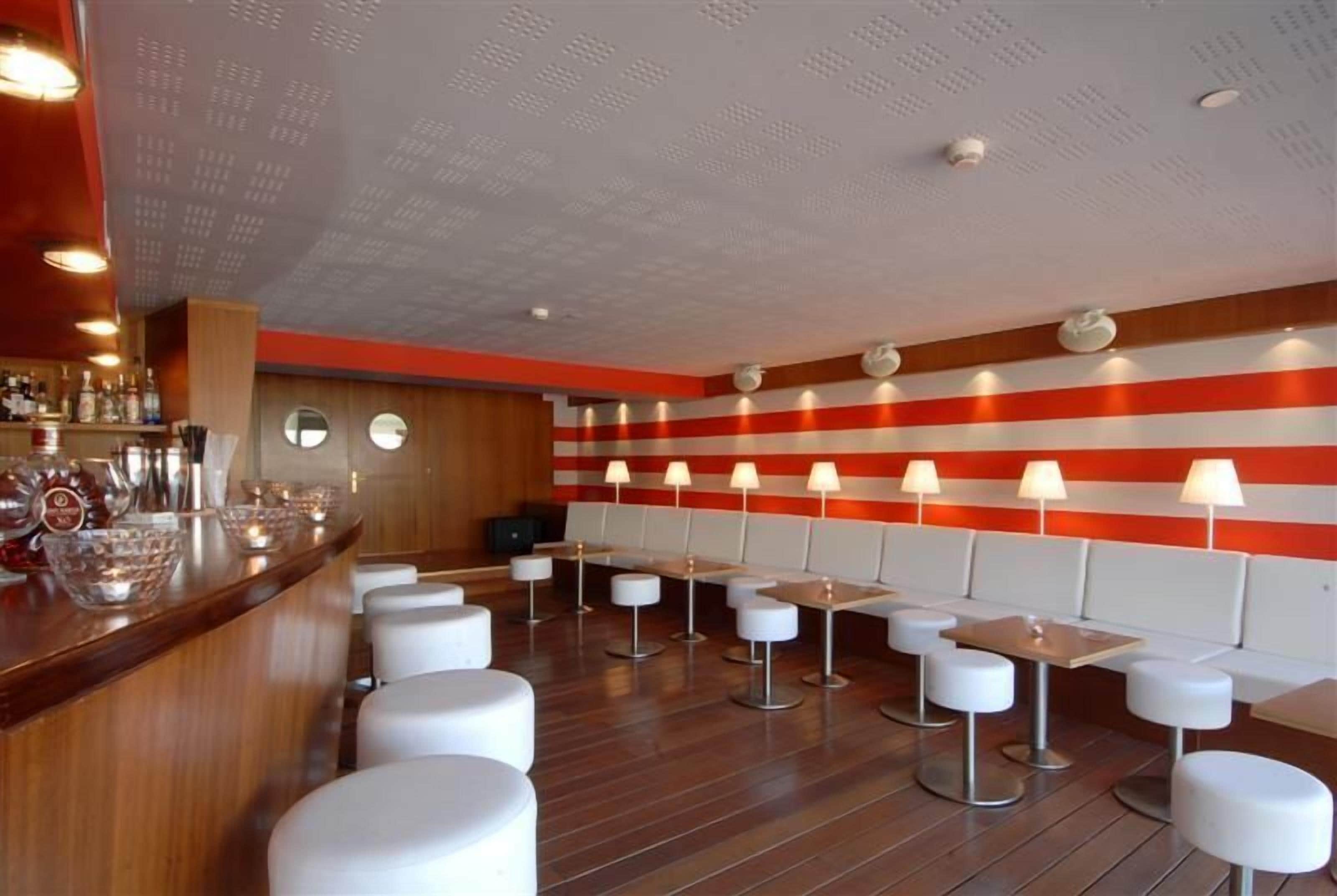 Una zona de bar con mostrador de madera, taburetes blancos y mesas con lámparas.