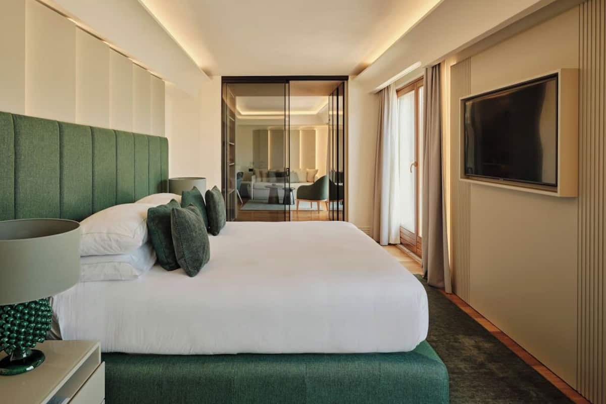 A hotel room with a bed, a TV, a desk with a lamp, and a view of the hallway.