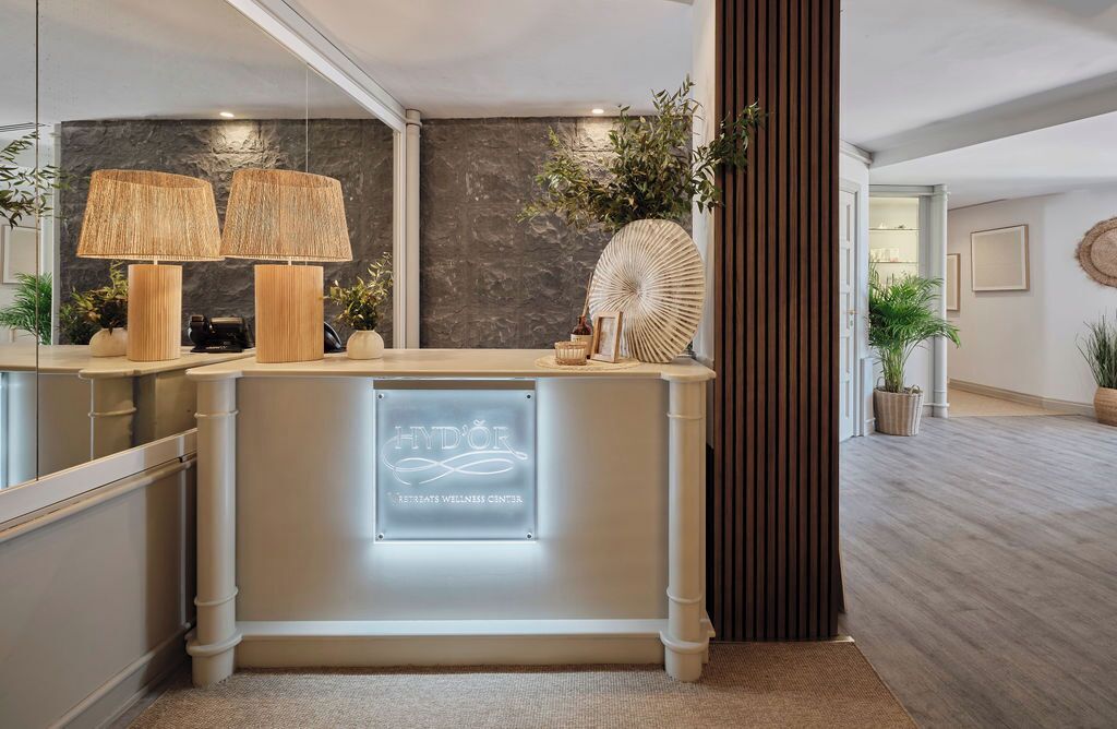 A reception desk with a sign reading 'HYD'OR Retreats Wellness Center,' two lamps, a decorative fan, and potted plants.