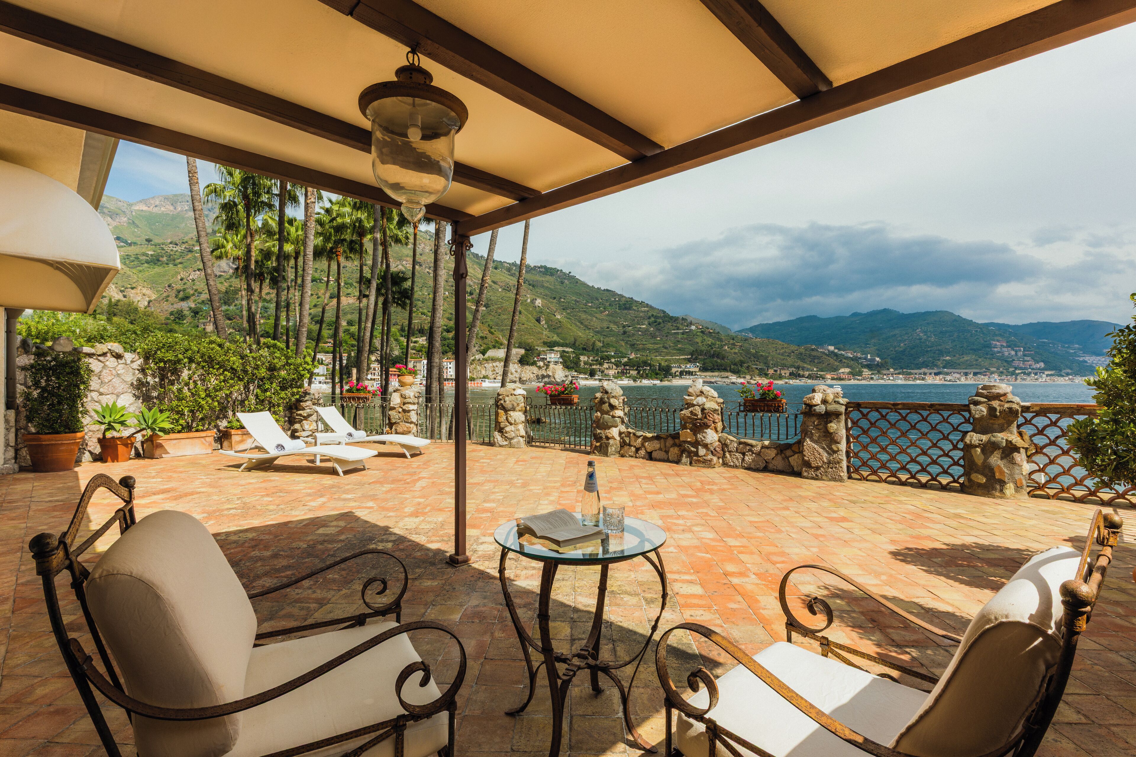 Une terrasse avec des meubles d’extérieur, une table avec un livre et un verre, et une vue sur la mer et les montagnes.