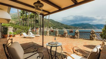 Um terraço com mobiliário externo, uma mesa com um livro e um copo, e vista para o mar e montanhas.