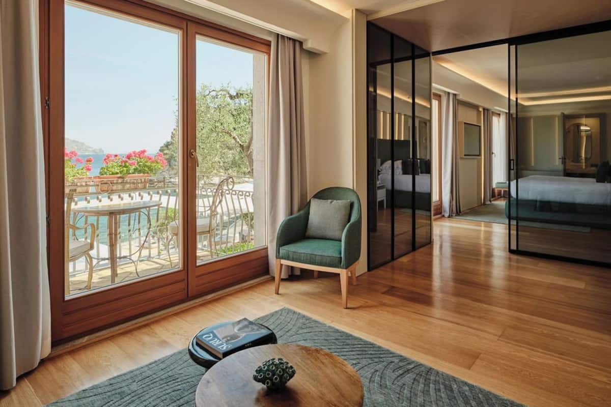 A modern hotel room with a large sliding glass door to a balcony, a green armchair, a bed with a metal frame, and a wooden floor.