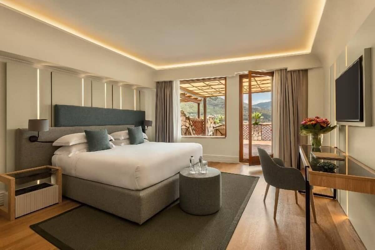 Quarto de hotel moderno com uma cama grande, uma escrivaninha com cadeira, televisão e varanda com vista.