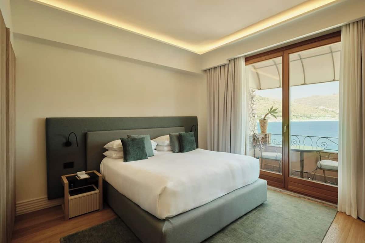 A hotel room with a large bed, a nightstand, a small table, and a balcony with a view.