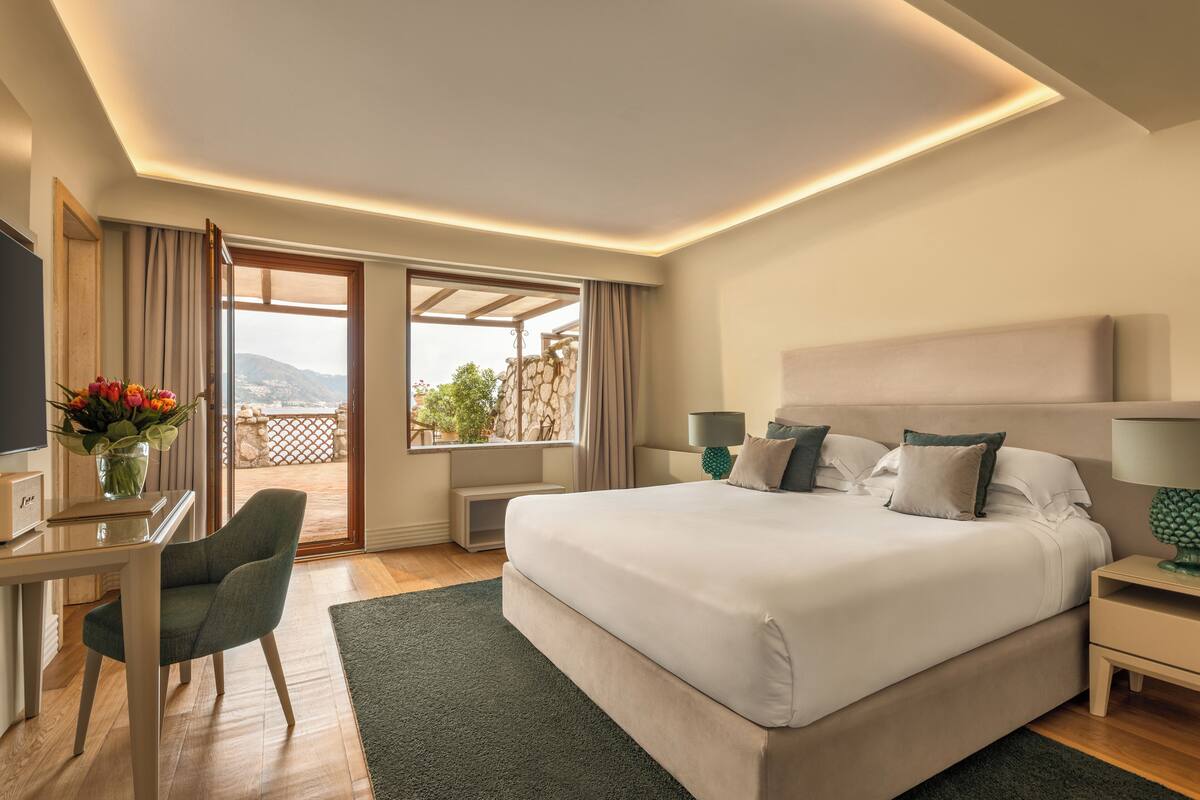 A modern bedroom with a large bed, a desk, and a view of the outdoors.