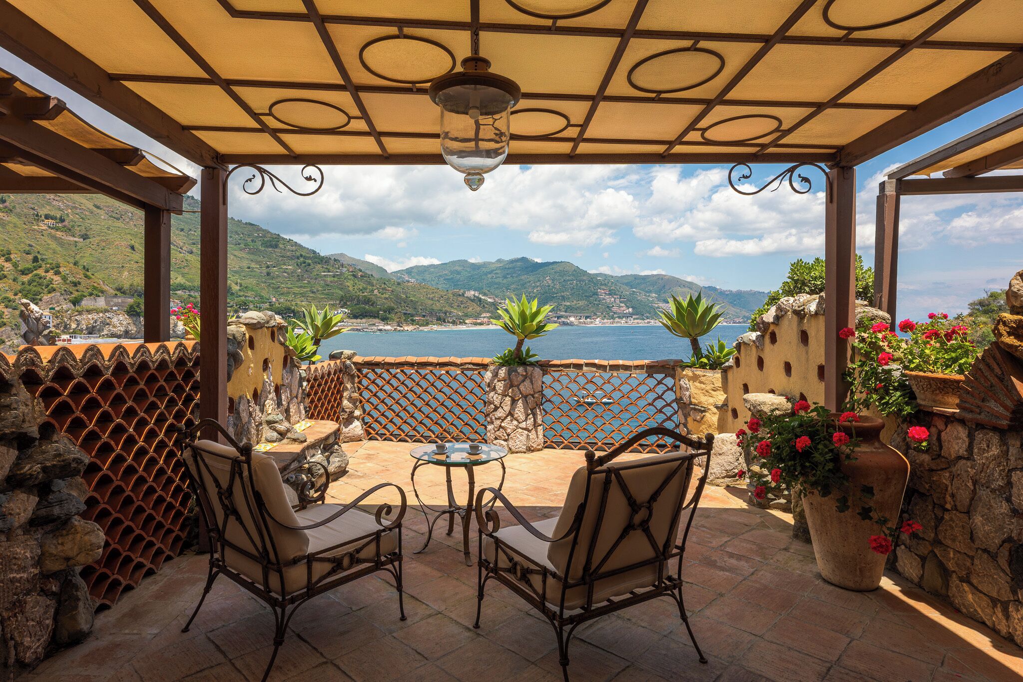 Überdachte Terrasse mit zwei Stühlen und einem Tisch, mit Blick auf eine malerische Küstenlandschaft mit Bergen und dem Meer.