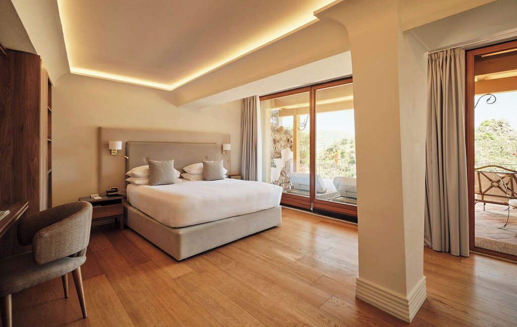 Une chambre spacieuse avec un grand lit, des tables de chevet, un fauteuil et une vue sur l’extérieur grâce à une porte-fenêtre coulissante.
