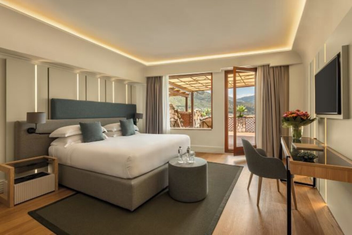 A modern hotel room with a large bed, bedside tables, a desk with a chair, a TV, and a balcony with a view.