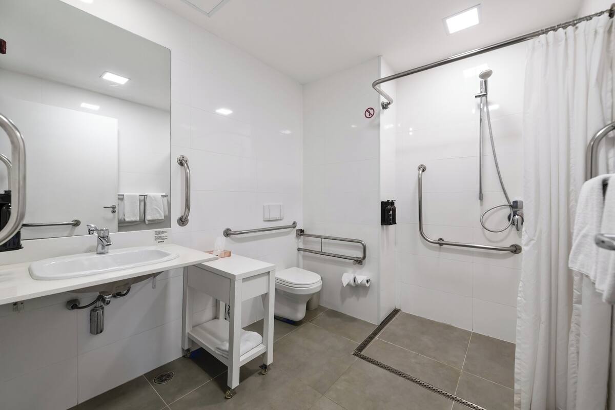 Une salle de bain moderne équipée d’un lavabo, de toilettes et d’une douche. Elle comprend également des barres d’appui et un sèche-mains.