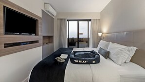Una habitación de hotel moderna con un televisor de pantalla plana, una cama cómoda con un cojín que dice "TO RELAX", una mesita de noche pequeña con un cuenco y un libro, y vistas de la ciudad a través de amplias ventanas.