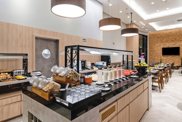 Un bar à petit-déjeuner moderne, avec des récipients en verre contenant des aliments, un espace dédié au café et un coin salon avec des tables et des chaises en bois.