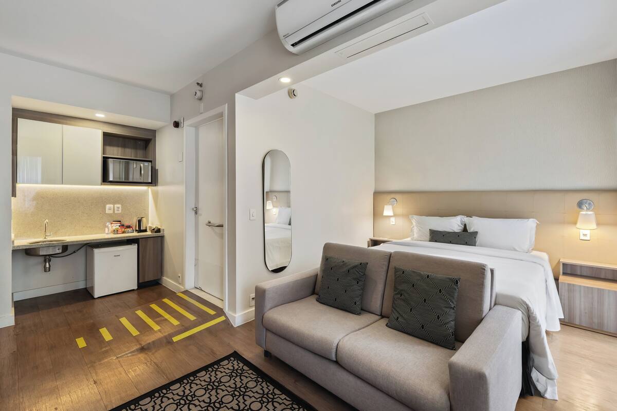 Une chambre d’hôtel moderne équipée d’une kitchenette, d’un canapé, d’un lit avec du linge de maison blanc et d’un grand miroir.