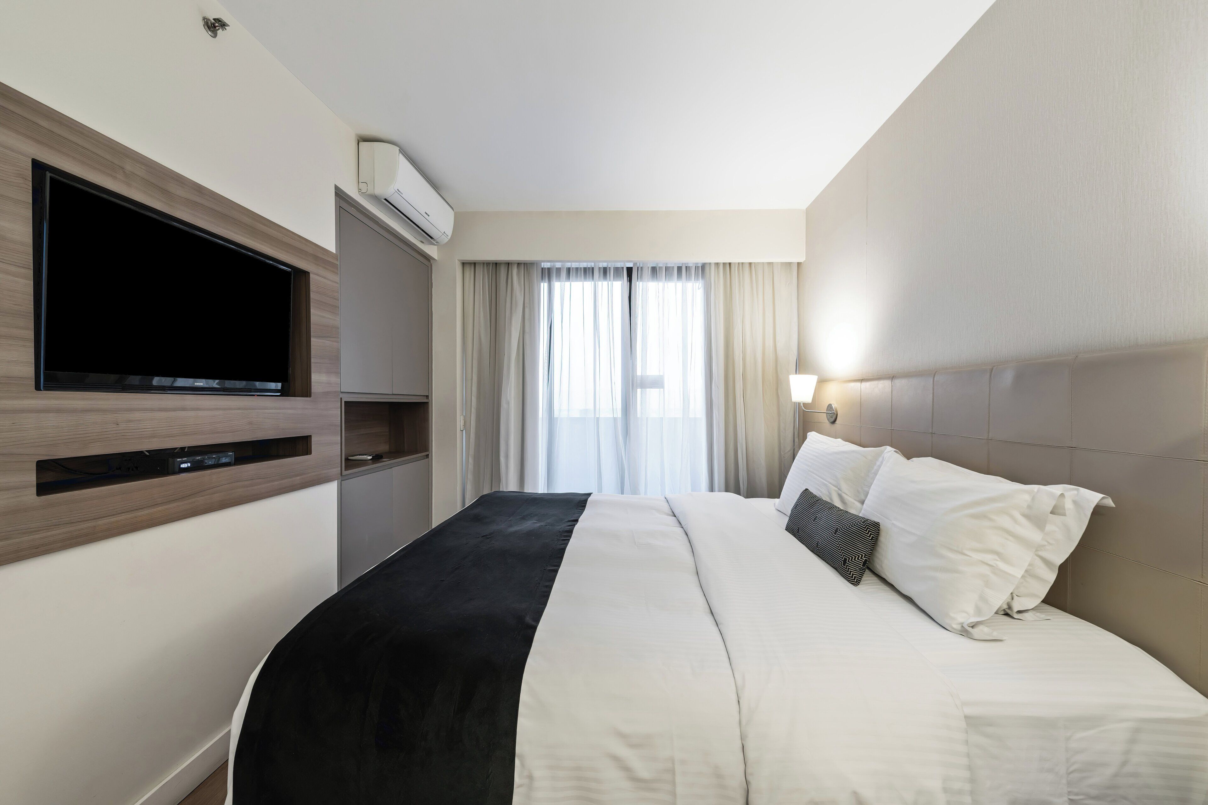Ett hotellrum med en stor säng, en platt-TV som är monterad på väggen, ett nattduksbord och ett fönster med gardiner.