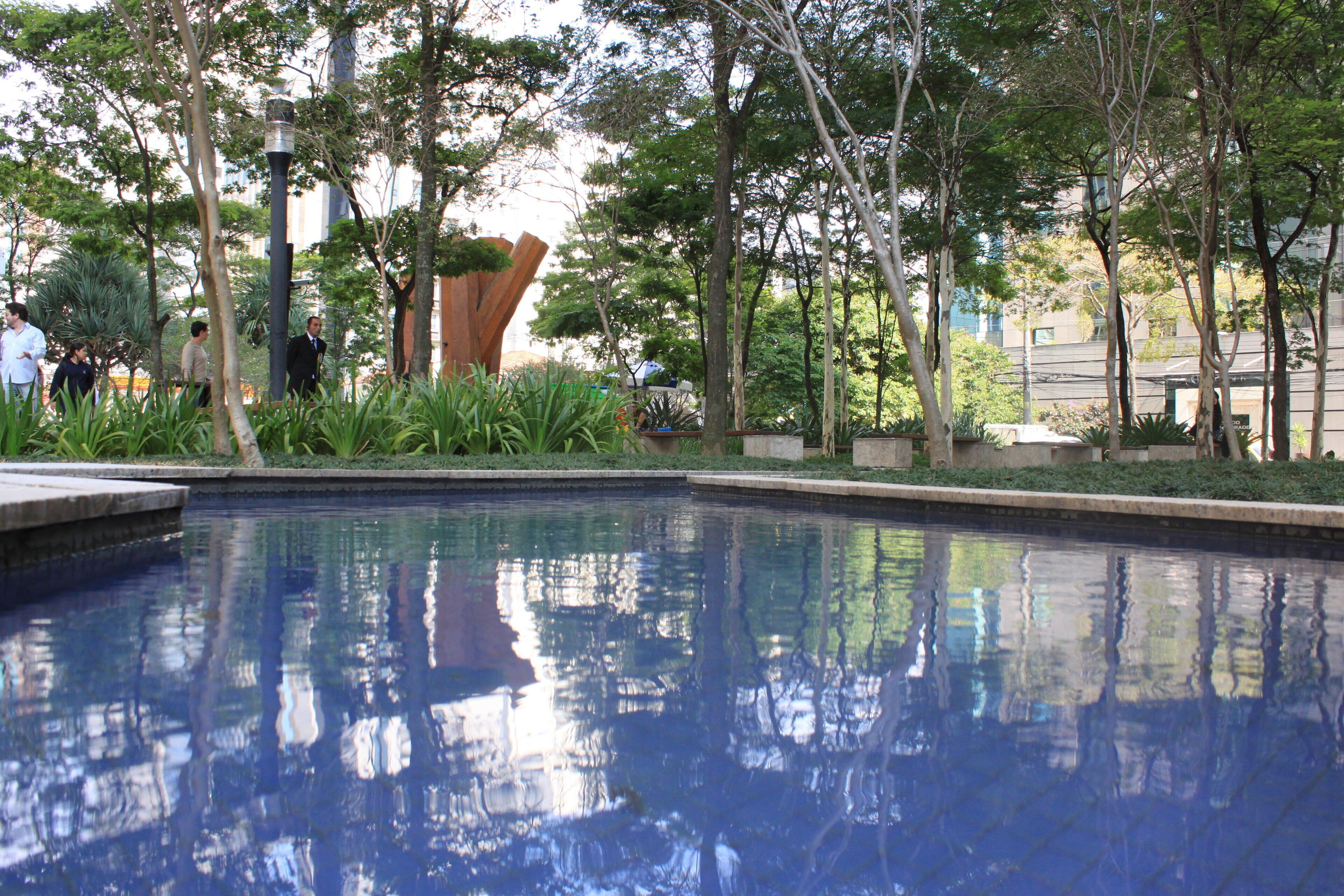 A swimming pool with a clear reflection of trees and buildings, surrounded by greenery and a modern building structure.